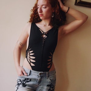 Peut inclure: Une femme portant un body noir avec des découpes et un détail de laçage à l'avant. Elle porte également un jean bleu clair avec des broderies noires.