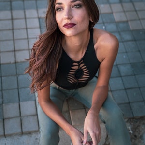 May include: A woman wearing a black crop top with a cut-out design and light green pants. She is sitting on a tiled surface and looking up.