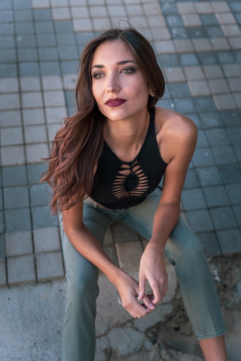 May include: A woman wearing a black crop top with a cut-out design and light green pants. She is sitting on a tiled surface and looking up.