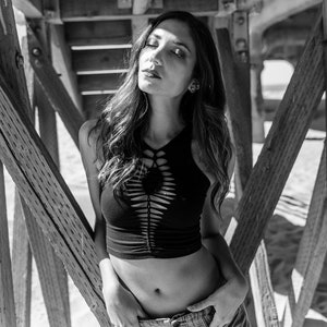 May include: A woman wearing a black crop top with cutouts and blue jeans stands under a wooden structure. The top has a criss-cross design and the jeans have a relaxed fit.