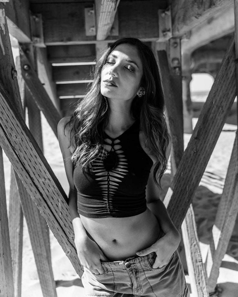 May include: A woman wearing a black crop top with cutouts and blue jeans stands under a wooden structure. The top has a criss-cross design and the jeans have a relaxed fit.