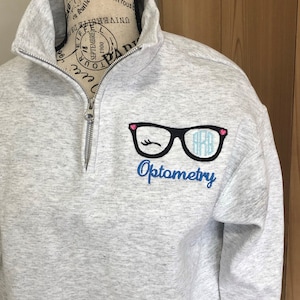 May include: A light gray quarter-zip sweatshirt with a black and pink glasses design and the word "Optometry" embroidered in blue. The glasses have the initials "ARB" inside. The sweatshirt is displayed on a mannequin.