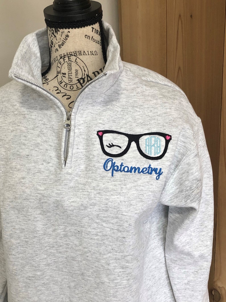May include: A light gray quarter-zip sweatshirt with a black and pink glasses design and the word "Optometry" embroidered in blue. The glasses have the initials "ARB" inside. The sweatshirt is displayed on a mannequin.