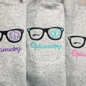 May include: Three gray hoodies with embroidered eyeglasses designs. Each pair of glasses has a different monogram and color accents, with the word "Optometry" below in matching colors. The glasses have eyelashes and heart details.