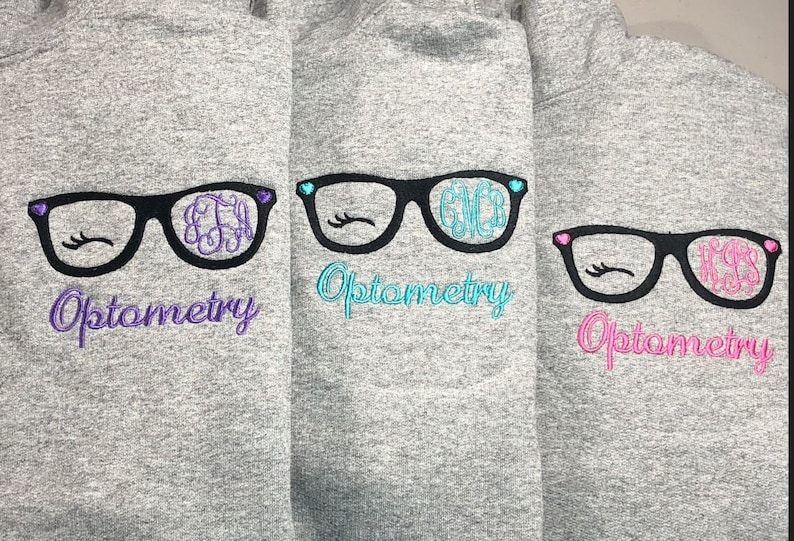 May include: Three gray hoodies with embroidered eyeglasses designs. Each pair of glasses has a different monogram and color accents, with the word "Optometry" below in matching colors. The glasses have eyelashes and heart details.