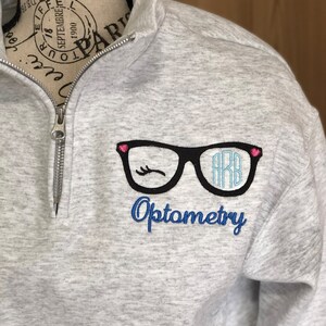 May include: A light gray Optometry sweatshirt with a quarter-zip neckline. The design features black eyeglasses with a blue monogram and a winking eye, and the word "Optometry" in blue script.