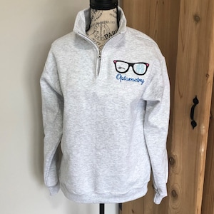 May include: A light gray quarter-zip sweatshirt with the word "Optometry" in blue script below a graphic of eyeglasses with a winking eye and initials. The sweatshirt has a high collar and a black zipper.