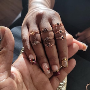 May include: A hand displays several copper wire-wrapped rings, each featuring a different gemstone. The rings are various sizes and designs, with stones in shades of yellow, purple, and brown. The nails are painted with a decorative design.