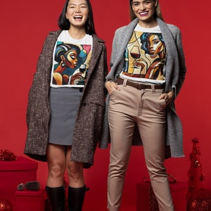 May include: Two people wearing white t-shirts with a colorful abstract design of a woman holding a glass of wine. The person on the left is wearing a brown tweed coat and a gray skirt. The person on the right is wearing a gray cardigan and beige pants. Both are wearing black boots. The background is a solid red.