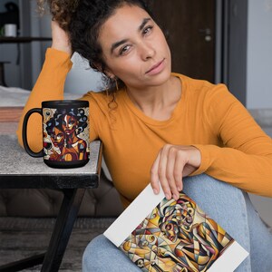 May include: A black coffee mug with a colorful illustration of two women with geometric patterns. The mug is on a black table. A person is holding a white notebook with a similar colorful illustration of a woman with geometric patterns.