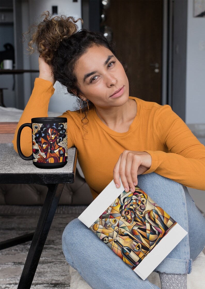 May include: A black coffee mug with a colorful illustration of two women with geometric patterns. The mug is on a black table. A person is holding a white notebook with a similar colorful illustration of a woman with geometric patterns.