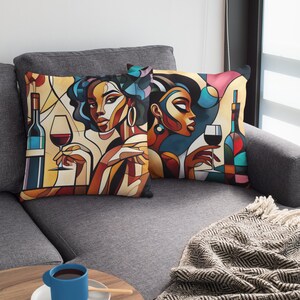 May include: Two colorful throw pillows with abstract art depicting women holding glasses of wine. The pillows are on a gray couch with a black and white throw blanket.