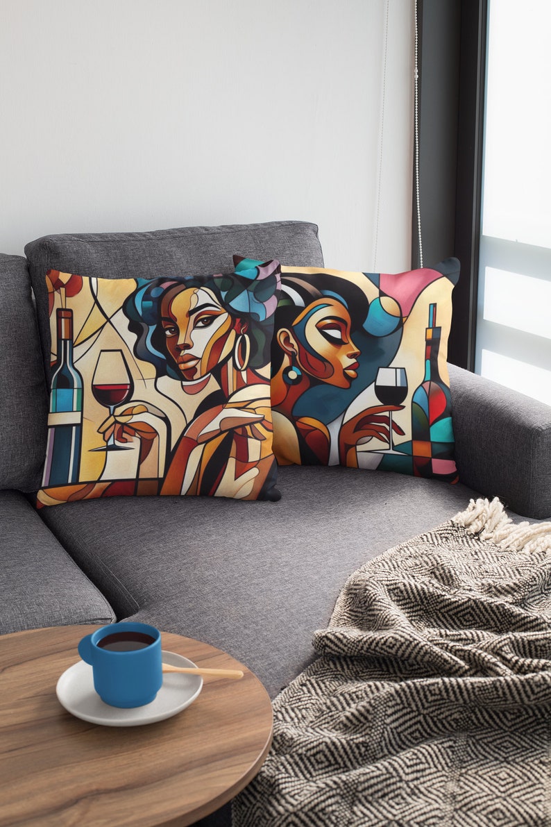 May include: Two colorful throw pillows with abstract art depicting women holding glasses of wine. The pillows are on a gray couch with a black and white throw blanket.