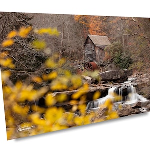May include: A scenic photograph of a wooden grist mill nestled beside a waterfall. The mill, with its weathered wood and water wheel, is surrounded by autumn foliage in shades of yellow, orange, and brown. The water cascades over rocks.