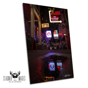 May include: A night photograph of a narrow street lined with glowing neon signs, including The Embers and The Climax Saloon, with their vibrant red, white, and blue lights reflecting clearly in a dark puddle on the ground in the foreground.