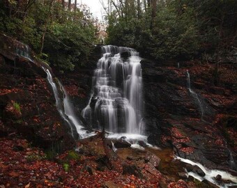 Peut inclure: Une cascade à plusieurs niveaux s'écoule sur des parois rocheuses sombres et humides, entourée d'un sol forestier dense jonché de feuilles d'automne. L'eau coule en filets blancs flous vers un petit bassin à la base, bordé d'arbres sombres.