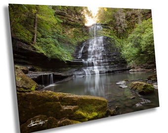 Peut inclure: Une impression murale rectangulaire représentant une cascade qui s'écoule sur des formations rocheuses sombres et stratifiées dans un bassin calme. La scène est entourée d'arbres verts luxuriants et de rochers moussus, avec une lumière vive.