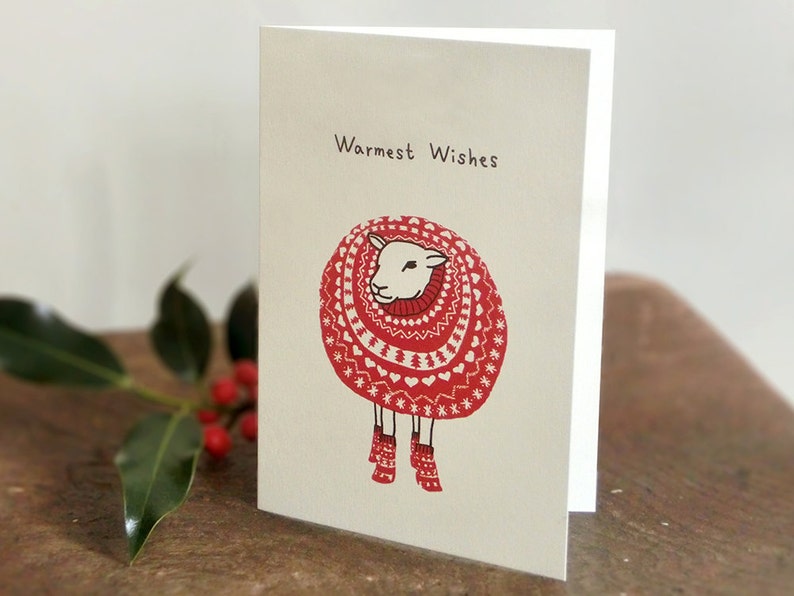 May include: A white sheep wearing a red and white knitted sweater with a heart pattern and red socks. The card says "Warmest Wishes".