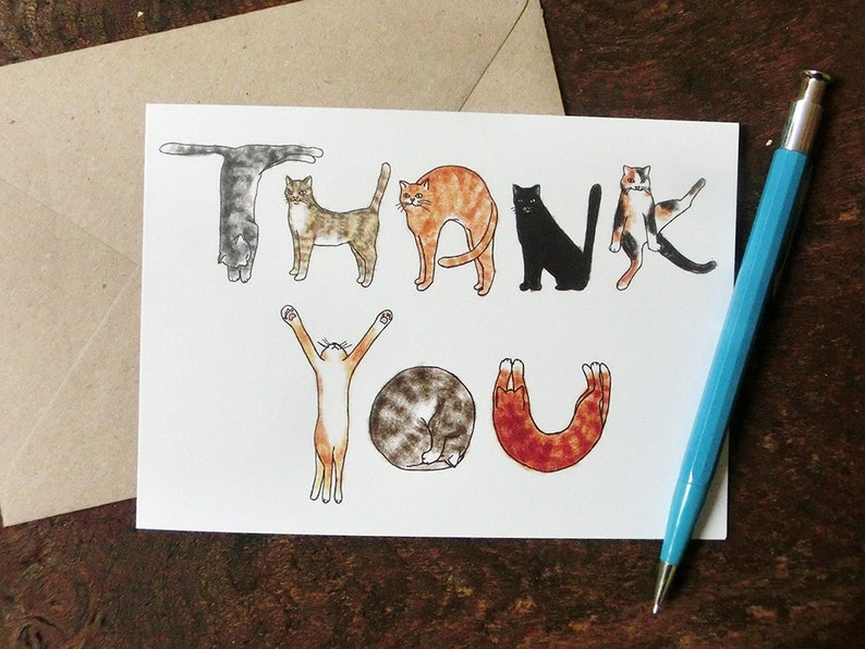 Pu&ograve; includere: Un biglietto bianco con la parola "GRAZIE" scritta usando illustrazioni di gatti in varie pose. I gatti sono disegnati in uno stile cartone animato e sono di diversi colori, tra cui nero, bianco, arancione e marrone.