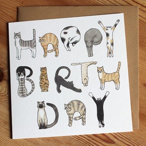 May include: A white card with a colorful illustration of cats forming the words "Happy Birthday". The cats are in various poses, including sitting, standing, and lying down. The card has a brown envelope.