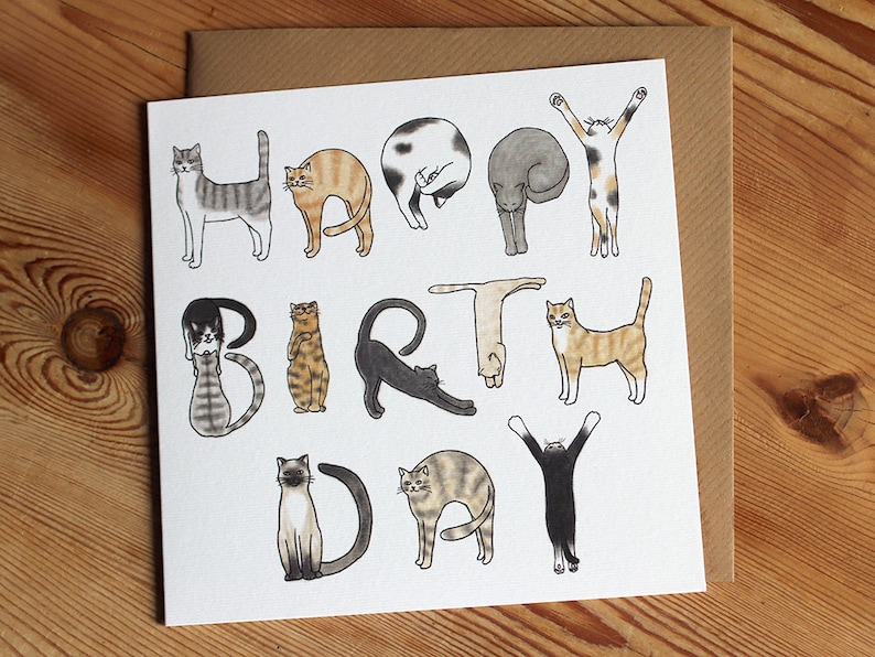 May include: A white card with a colorful illustration of cats forming the words "Happy Birthday". The cats are in various poses, including sitting, standing, and lying down. The card has a brown envelope.