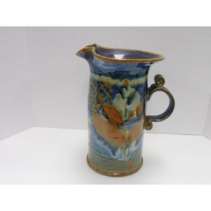 May include: A tall, ceramic pitcher with a unique, organic shape. The pitcher features a dark blue rim and handle, with a body adorned with blue, brown, and cream-colored abstract patterns. The handle has a decorative swirl design.