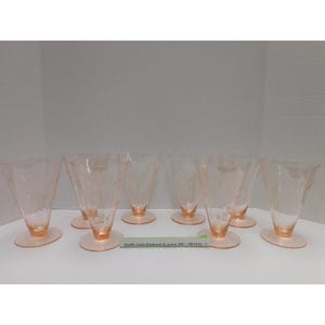 May include: A set of seven light pink glass tumblers with a flared, conical shape and a circular base. Each glass features a delicate etched floral pattern. A small green rectangular label with the text North coast diamond & pawn 330-749-0141 is placed in front of the glasses.