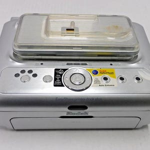 May include: Silver Kodak EasyShare printer dock plus with a clear plastic top. Features several buttons and a central dial. Includes Kodak Perfect Touch and Auto Enhance labels. The device has a small, rectangular opening at the bottom.