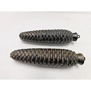 May include: Two antique clock weights shaped like pine cones. The weights are dark brown with a textured surface, and have a metal loop at the top. The weights are approximately 5 inches long.