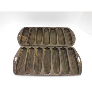 May include: Two cast iron corn-shaped baking pans. Each pan has seven corn cob molds. The pans are dark brown with a rustic appearance, featuring handles on each end. Ideal for baking cornbread.