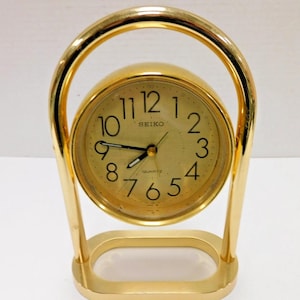 May include: A gold-toned Seiko quartz clock with a round face and a gold arched handle. The clock face has black numerals and hands. The clock is supported by a rectangular base.