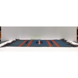 May include: A rectangular woven textile runner in a muted blue hue featuring orange and red vertical stripes. The center displays a geometric motif in black, red, and white. The short ends are finished with dark, fringed edges on both sides.