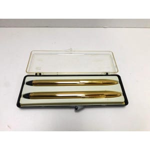 May include: A set of two gold-toned pens with black accents, housed in a rectangular black plastic case with a clear, hinged lid. The pens feature a smooth, metallic finish and are positioned horizontally within a molded white interior tray.