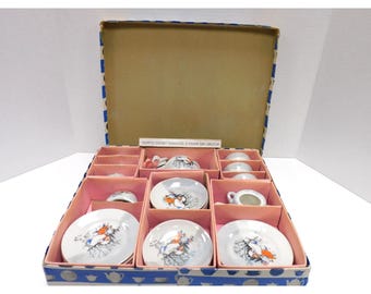 May include: A vintage miniature tea set in a box. The set includes small white plates, cups, and a creamer, each decorated with a bird and branch design in orange, blue, and black. The box is blue and pink.