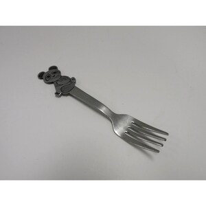 May include: A stainless steel fork with a decorative koala bear design at the top. The fork has four tines and a long handle. The koala is in a sitting position.