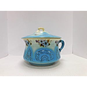 May include: A vintage ceramic container with a lid, featuring a light blue and cream color scheme. The container is adorned with floral and seashell designs, and has a handle. The lid has a small handle on top.