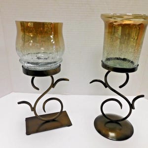 May include: Two decorative candle holders. Each features a clear glass cup with a gold-colored rim, set on a dark bronze metal stand. The stands have a swirling, artistic design, with one on a rectangular base and the other on a circular base.