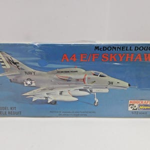 May include: A model kit box for a McDonnell Douglas A4 E/F Skyhawk aircraft. The box features a detailed illustration of the plane in flight, with "NAVY" and "311" visible. The box also includes the text "MODEL KIT MODELE REDUIT" and "1/72 SCALE".