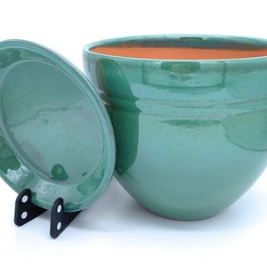 May include: A teal glazed ceramic planter with a matching saucer. The planter has a rounded shape and a slightly flared rim.