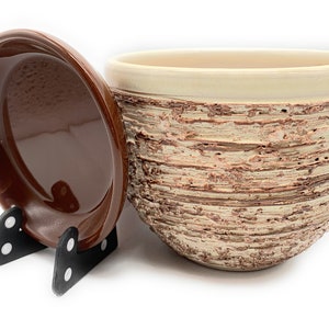 May include: A ceramic planter with a textured, bark-like exterior in beige and brown tones. The top rim is a solid beige colour. A matching brown saucer sits on a black stand, next to the planter. Suitable for indoor or outdoor use.