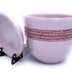 May include: A pink ceramic container with a lid, featuring a decorative band of brown textured lines around the middle. The container is sitting on a white surface.
