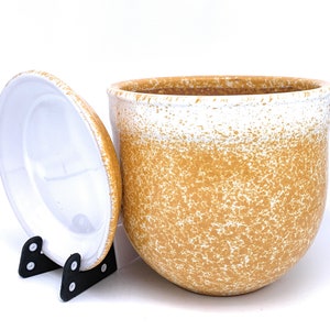 May include: A white ceramic planter with a speckled brown glaze. The planter has a rounded bottom and a wide opening. The planter is shown with its lid off, which is also white with a speckled brown glaze.