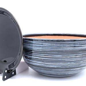 May include: A black ceramic bowl with a matching lid, both featuring a horizontal white stripe design. The bowl has a wide opening and a terracotta-coloured interior. The lid is propped up on a black stand.