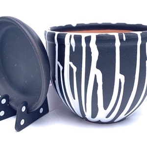 May include: Black and white striped ceramic planter with a matching lid. The planter has a rounded shape and a white rim. The lid is flat and has a black rim.