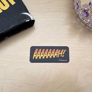 Puede incluir: Una pegatina rectangular con fondo oscuro que presenta el texto amarillo y naranja AAAAAAAH! y el texto más pequeño -Prepotente en blanco. La pegatina está colocada cerca de un objeto negro con texto amarillo y una corona plateada con gemas moradas.
