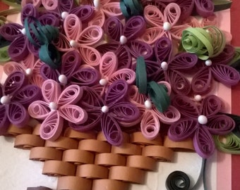 May include: A handmade paper flower bouquet in shades of pink and purple, with green leaves and a brown basket. The flowers are made using the quilling technique, where paper is rolled and shaped to create intricate designs.