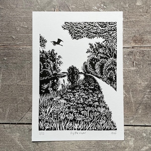 May include: A black and white linocut print titled "By the river." The artwork shows a river scene with a path, trees, and a flying bird. The print is numbered 2/25 and signed by the artist.