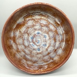 May include: A round ceramic bowl featuring a crystalline glaze with a repeating pattern of white, star-like bursts against a warm, reddish-brown background. The interior surface displays a textured, radial design with subtle blue accents at the center.