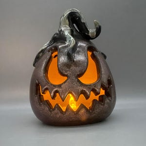 May include: A dark brown ceramic jack-o'-lantern with a glossy finish and a curved stem. The pumpkin features cut-out eyes and a jagged mouth, illuminated from within by a warm orange light that glows through the openings in the ceramic body.