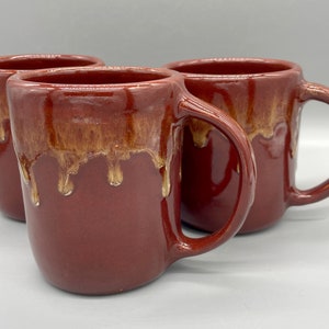 May include: Three red ceramic mugs with a drip glaze in a light brown color. The mugs have a rounded shape and a handle.
